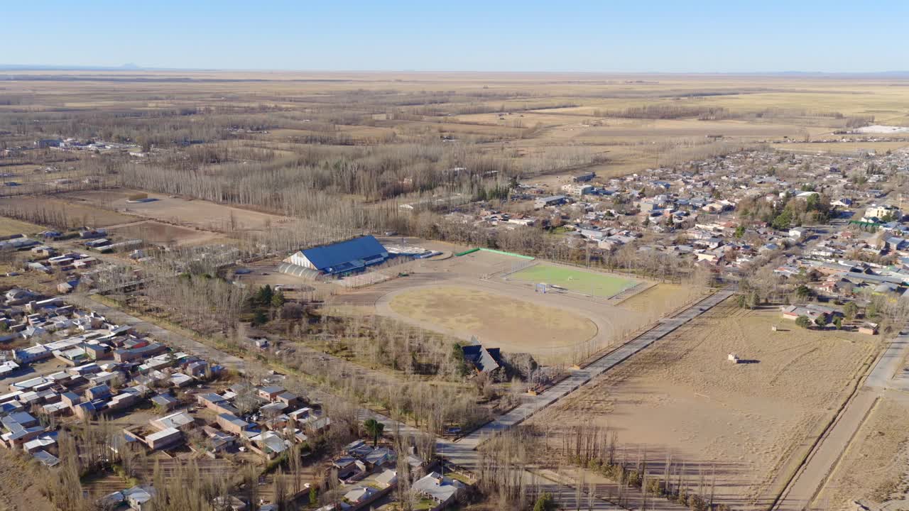 Drone shot over Polideportivo Municipal in Malargüe, Mendoza, showing sports facilities and surrounding dry landscape with farmland, houses, and autumn trees under clear skies