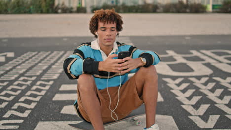 smiling guy enjoying music on phone outdoors closeup. hipster sitting skateboard