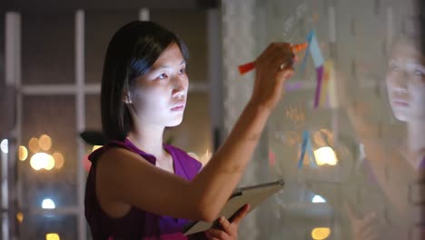 Asian-casual-businesswoman-using-tablet-brainstorming,-making-notes-on-glass-wall-at-night-in-office