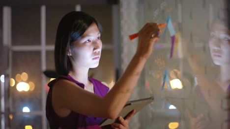 Mujer-De-Negocios-Asiática-Casual-Usando-Una-Lluvia-De-Ideas-En-Una-Tableta,-Tomando-Notas-En-Una-Pared-De-Vidrio-Por-La-Noche-En-La-Oficina