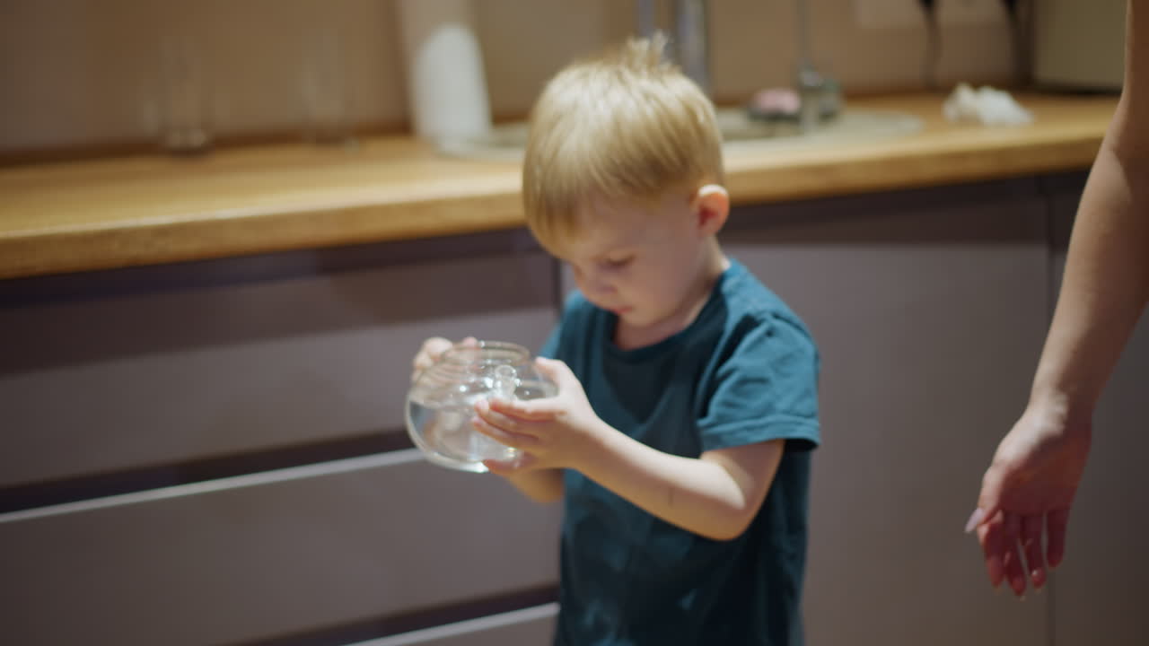 Child holding glass teapot filled with clear water near sink, close up view highlighting small hands, transparent container, clean liquid, everyday home moment, concept of purity, hydration, responsibility