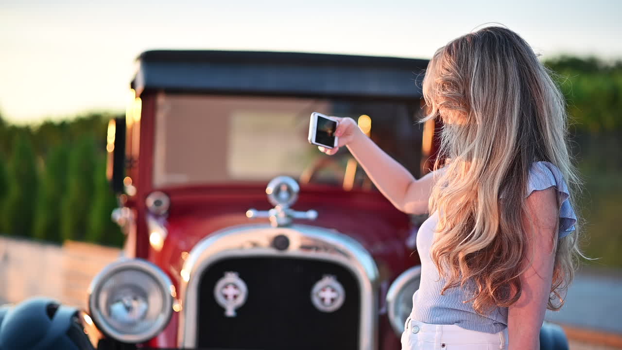 Young woman holding smartphone and taking a selfie near a red vintage car at sunset