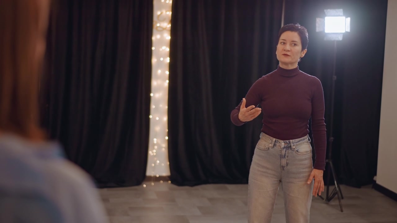 Female performer practices audition monologue under stage lights, receiving director feedback and hand gestures from coach curtain seam and string lights create warm backdrop confident delivery