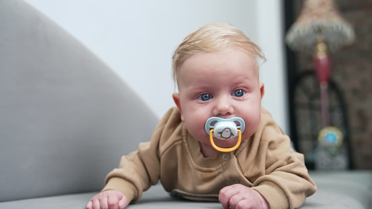 Lovely blond Caucasian baby boy with tears in big blue eyes. Infant child with pacifier lies on belly. Dad's hand wants to get a pacifier but kid turns away. Close up.