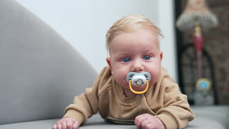 lovely blond caucasian baby boy with tears in big blue eyes. infant child with pacifier lies on belly. dad's hand wants to get a pacifier but kid turns away. close up.