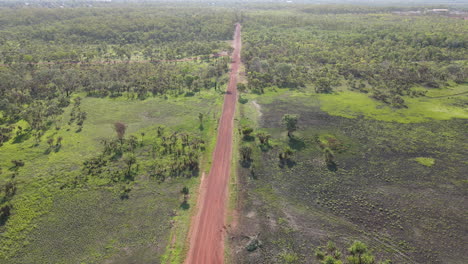 Toma-De-Un-Dron-De-Movimiento-Lento-Y-Alto-De-Una-Carretera-Roja-Recta-Vacía-Y-Matorrales-Verdes-Cerca-Del-Parque-Natural-De-La-Jungla-De-Holmes,-Darwin,-Territorio-Del-Norte