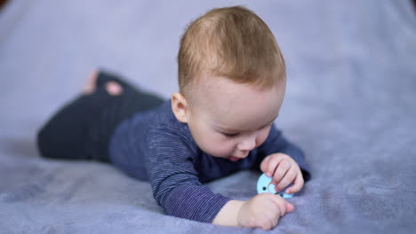 little baby boy resting on big bed lies on his belly. beautiful caucasian kid holds a pacifier in hands and looks at it attentively. close up.