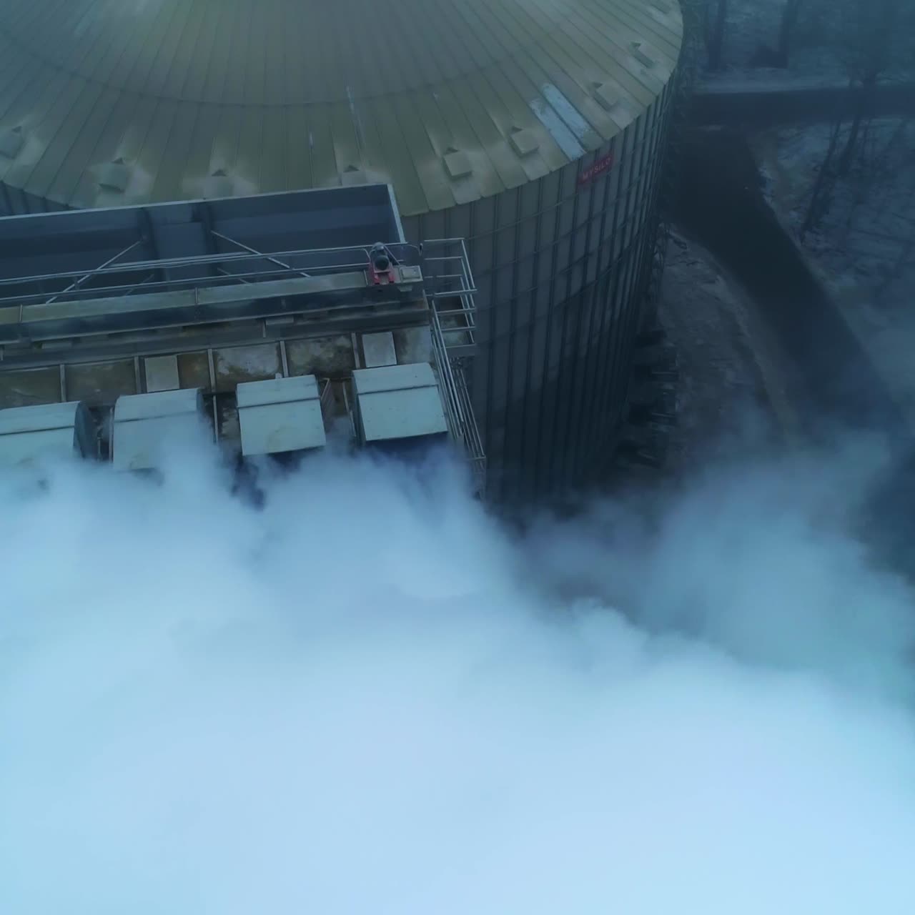 Five connected pipes producing heavy thick white smoke. Granary complex premises and tanks from aerial perspective on grey winter day