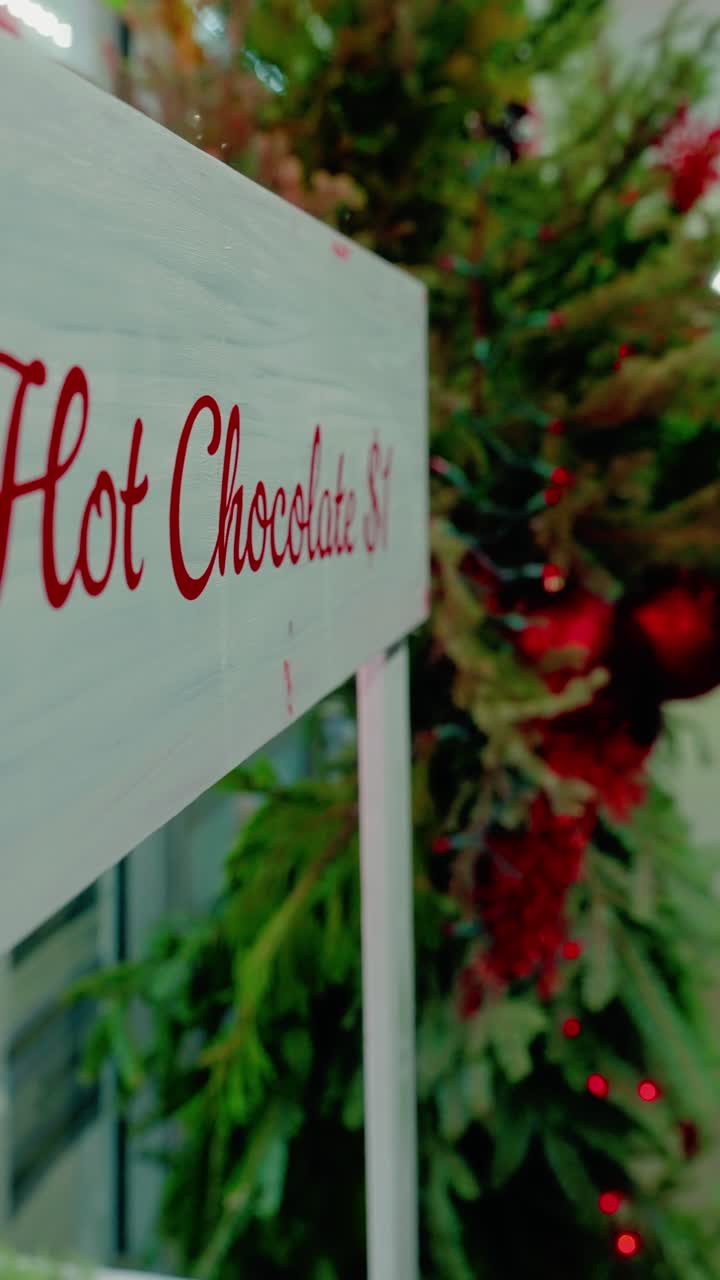 Cartello del prezzo della cioccolata calda bianca accanto a un albero sempreverde decorato con ornamenti rossi e luci, fuori da un negozio locale di notte.