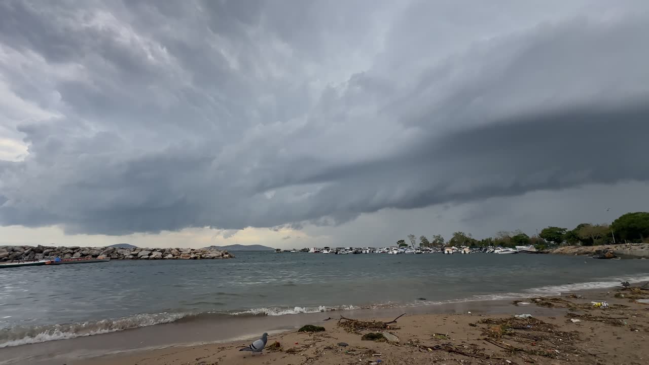 Dramatic dark storm clouds gather over a rough sea. Waves crash on the shore near a stone breakwater and a marina with boats on a gloomy autumn day