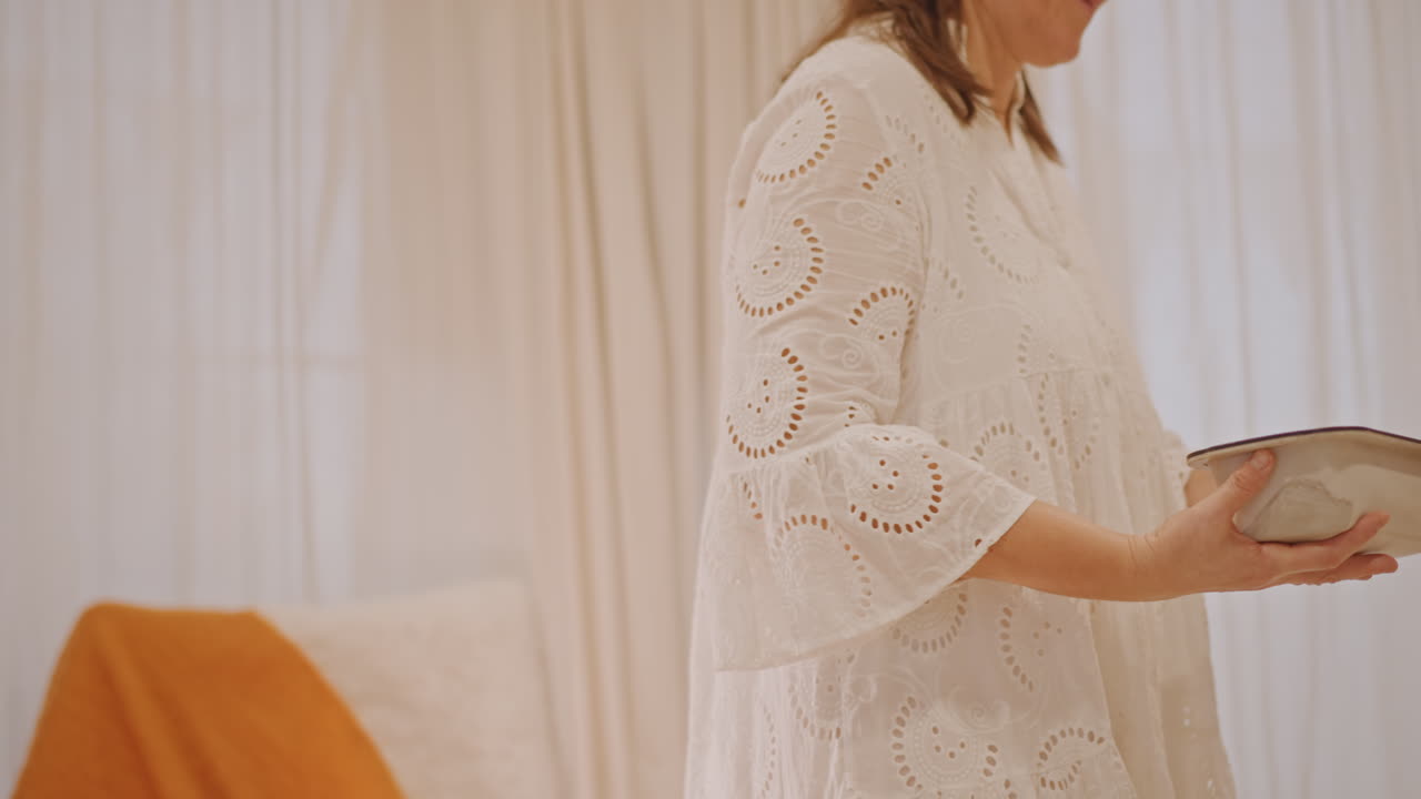 Eine weiße Frau trägt eine Kürbiskuchenform über einen warmen Küchentisch und schiebt sie dann in den Ofen, in einer gemütlichen Herbstbackszene. Dazu ein Spitzenkleid, ein oranges Plaid auf einem Stuhl, ein Teekessel auf dem Herd und sanftes Morgenlicht.