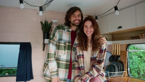 Portrait-of-a-happy-couple,-a-brunette-guy-with-stubble-in-a-green-checkered-shirt-hugs-his-brunette-girlfriend-in-a-pink-checkered-shirt-in-their-campervan-outside-the-city-in-a-camp-during-a-picnic-in-the-summer