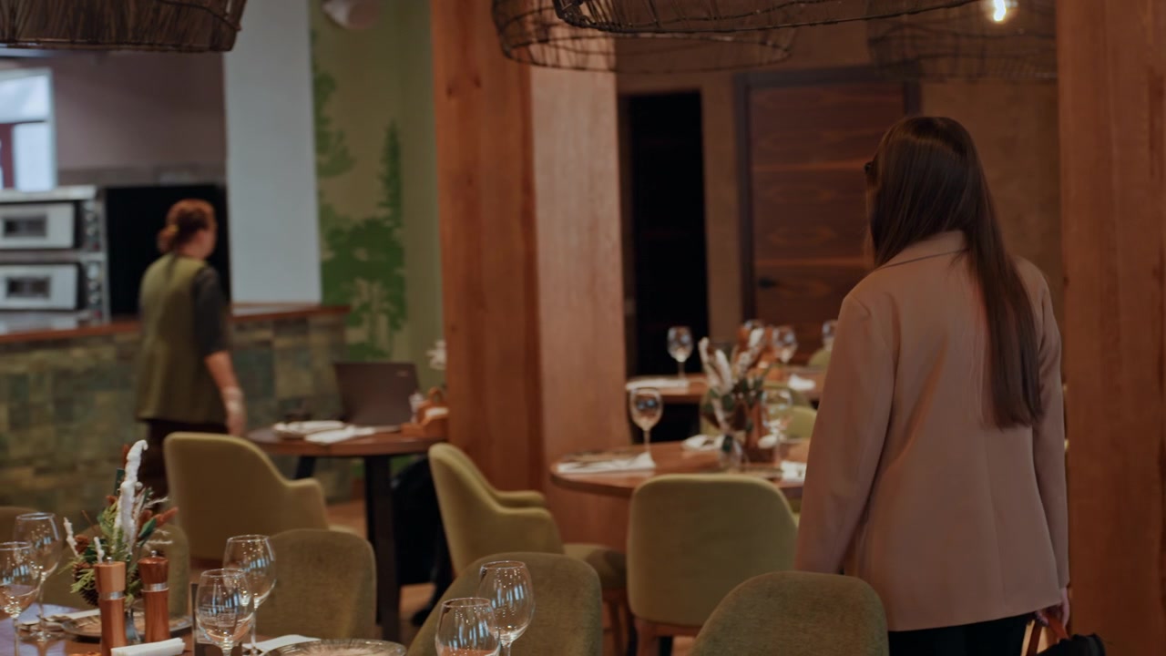 Donna in piedi nella sala da pranzo vuota di un ristorante, cammina lentamente tra tavoli apparecchiati e sedie verdi, colonne di legno caldo e illuminazione a sospensione soffusa, bicchieri e centrotavola sulle apparecchiature dei tavoli
