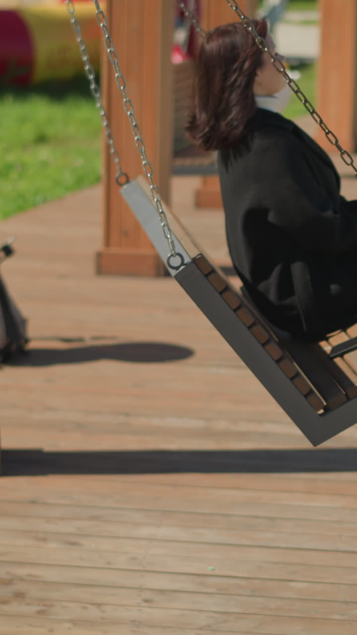 Side view of lady in stylish black coat and boots relaxing on wooden swing in sunlit park, surrounded by greenery and decorative flowers, under bright sky with wooden structures in background