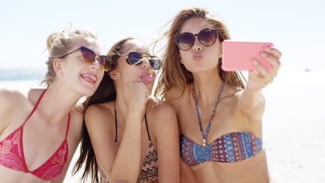 Three-teenage-girl-friends-taking-selfie-on-beach-pulling-funny-faces-wearing-colorful-bikini-sharing-vacation-photo