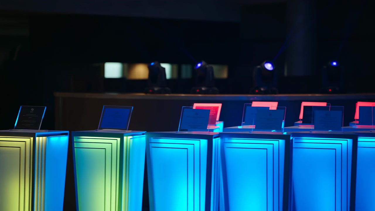 Low-angle interior view of illuminated exhibition pedestals lined in row, each topped with angled transparent display panels, set within dark modern venue with reflective surfaces and vivid colored