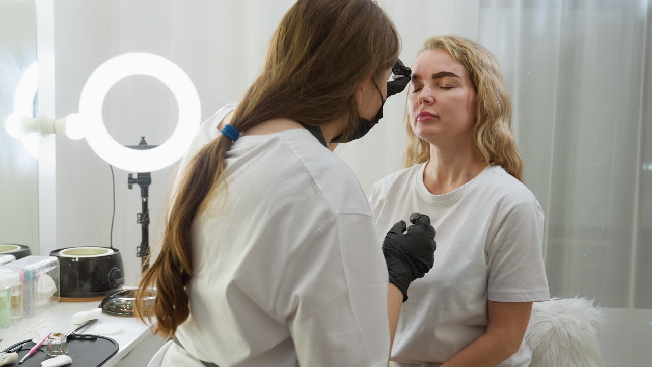 Beautician wearing gloves cleans eyebrow of seated client in bright salon using cotton pad, with professional tools, furry chair, ring light, and cosmetic items clearly visible in organized background