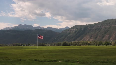 large american flag waving in the wind over a grassy field and cloudy sky with the san juan mountains in background