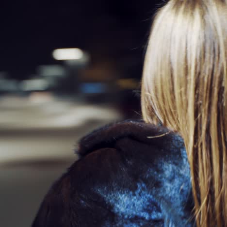 A joyful girl in a black half-length coat is holding a violin in her hands and playing on it in a winter evening in the square in the center of the city. Blurred background. Camera motion to around