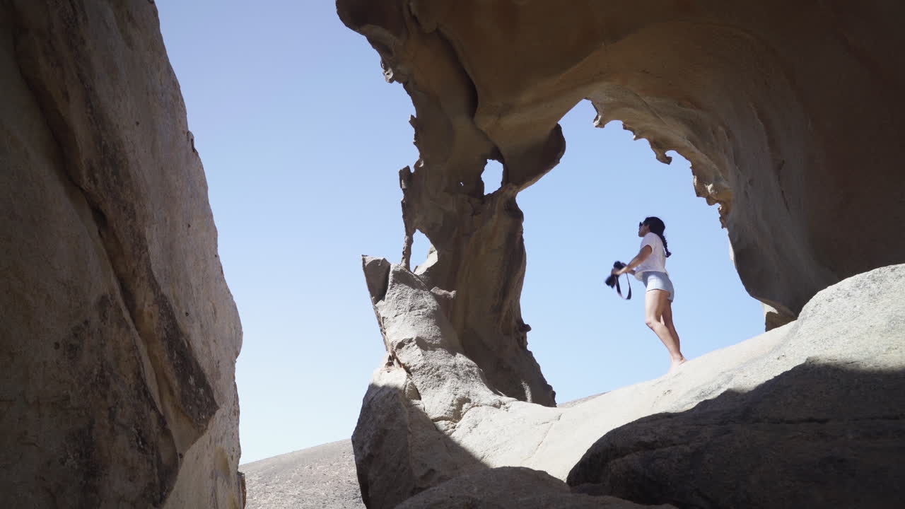 joven fotógrafa de pie dentro de la formación de arco rocoso en las montañas del desierto de fuerteventura isla canaria españa con cielo azul claro durante el día soleado