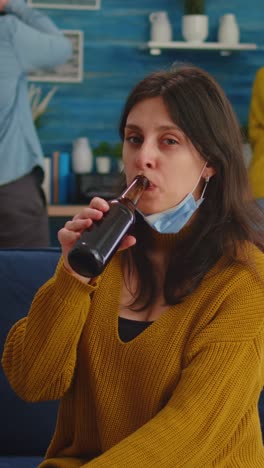 vertical video: young lady with protection mask in living room drinking beer