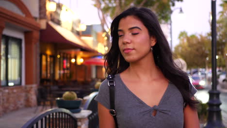 A-beautiful-hispanic-woman-walking-a-downtown-urban-city-street-looking-in-restaurants,-shops-and-retail-stores-with-lights-at-twilight-SLOW-MOTION