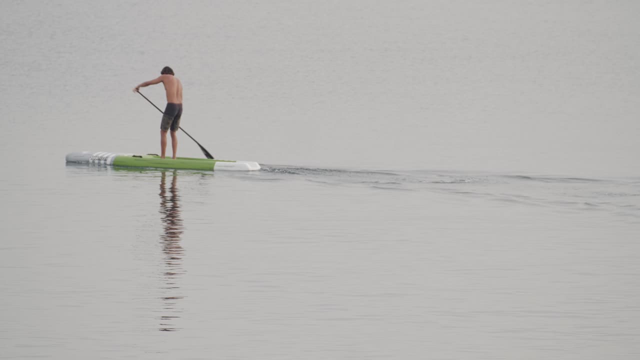 un joven en forma rema en una tabla de remo en un mar en calma al amanecer en cámara lenta de 60 fps