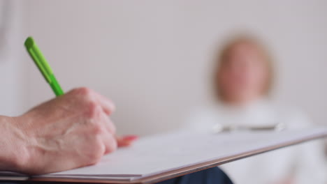female hand holding green pen noting on clipboard during counseling session, with client seated in background, slightly blurred, suggesting ongoing therapeutic exchange in calm setting