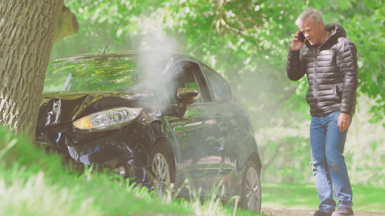 성숙한 남자가 담배를 피우는 차에서 나와 응급 서비스에 전화하기 전에 사고 피해를 검사합니다.