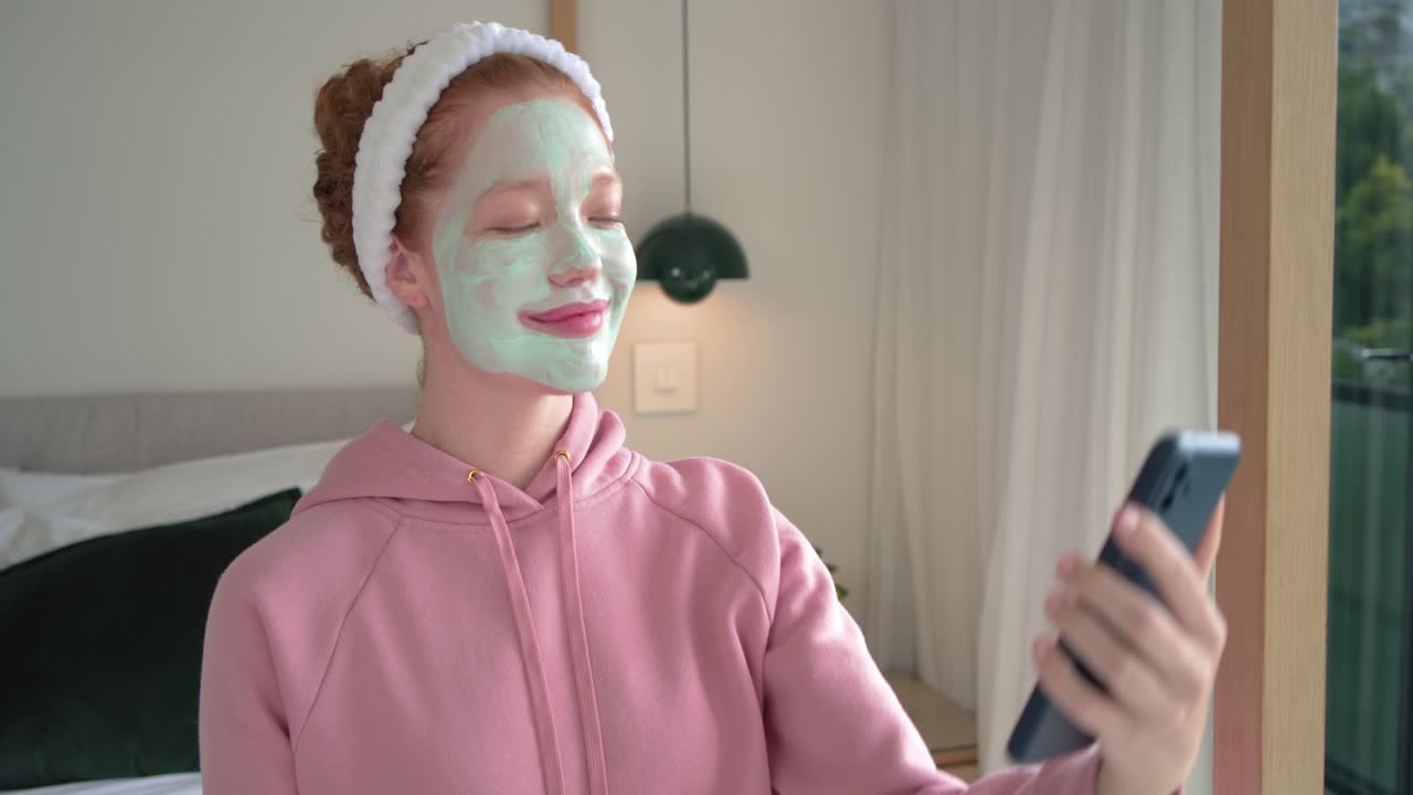 Red-haired woman sitting on bed in hoodie, scrolling phone then raising phone, posing mask selfie