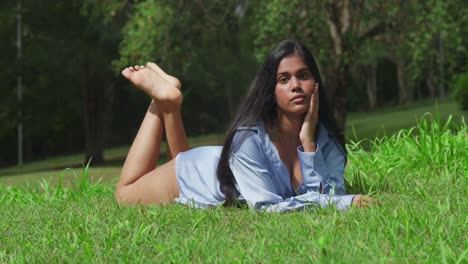 in a blue shirt, a young east indian girl at a park laying in the grass with her feet in the air