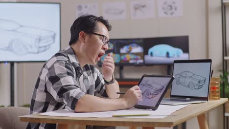 vista lateral de un hombre asiático bostezando y durmiendo mientras dibuja un nuevo concepto de diseño de automóvil en una tableta en el estudio con tv y computadoras muestra modelo de automóvil eléctrico 3d
