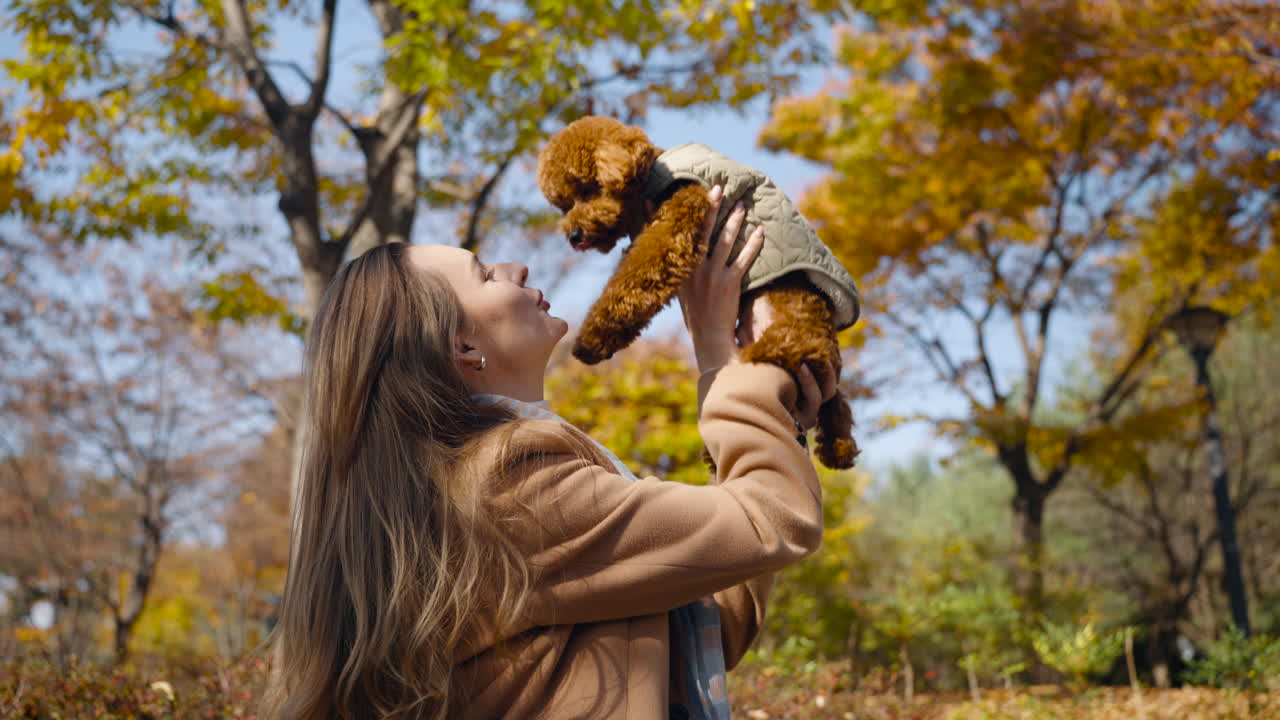 A cheerful woman joyfully lifted a small brown Toy Poodle dog in her arms up in front of her, kicking and caressing him while standing outdoors in a park with vibrant autumn foliage - slow motion