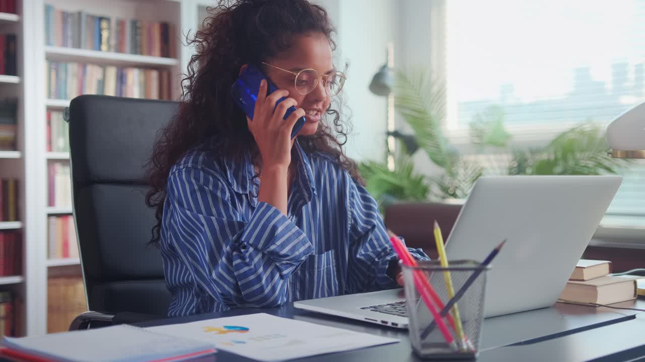Smilende indisk kvinde arbejder på bærbar computer fra hjemmekontor og taler i mobiltelefon.