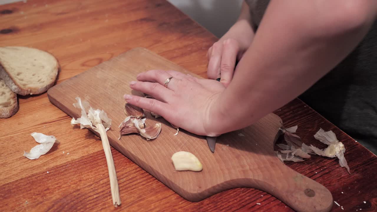 Woman's hands with a knife crush the garlic on a chopping board, then peel the skin-1