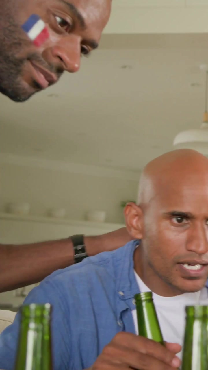 Vertical video: Sipping beer bald man in blue shirt calming during game at home with painted friend