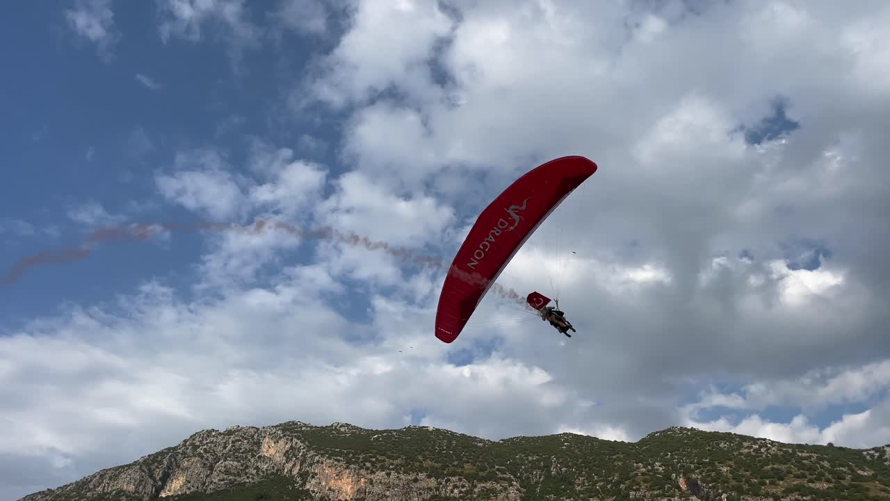 Turkey,Fethiye,Oludeniz,Footage starts with a red paragliding carrying a Turkish flag and with a smoke behind it,gliding slowly towards to the boardwalk with many people around. Beautiful Parade start