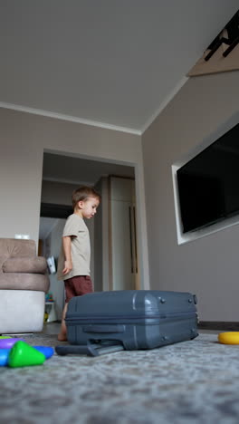 small baby boy comes up to a big suitcase and drops it on the floor. little kid plays with valise wheels. low angle view. vertical video.