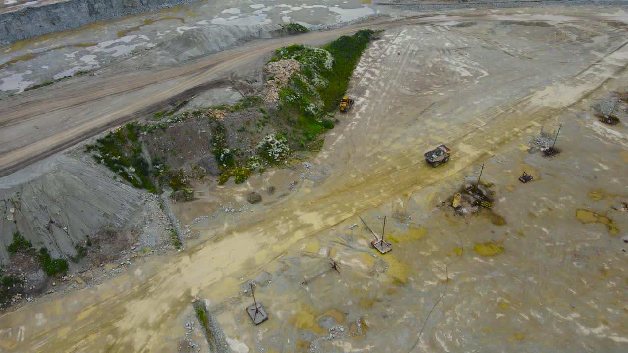 Top down aerial drone footage revealing a large tipper lorry heavy mining machine carrying rocks at a wet and damp limestone quarry during a cloudy day in a pit that has yellow puddles, gravel piles