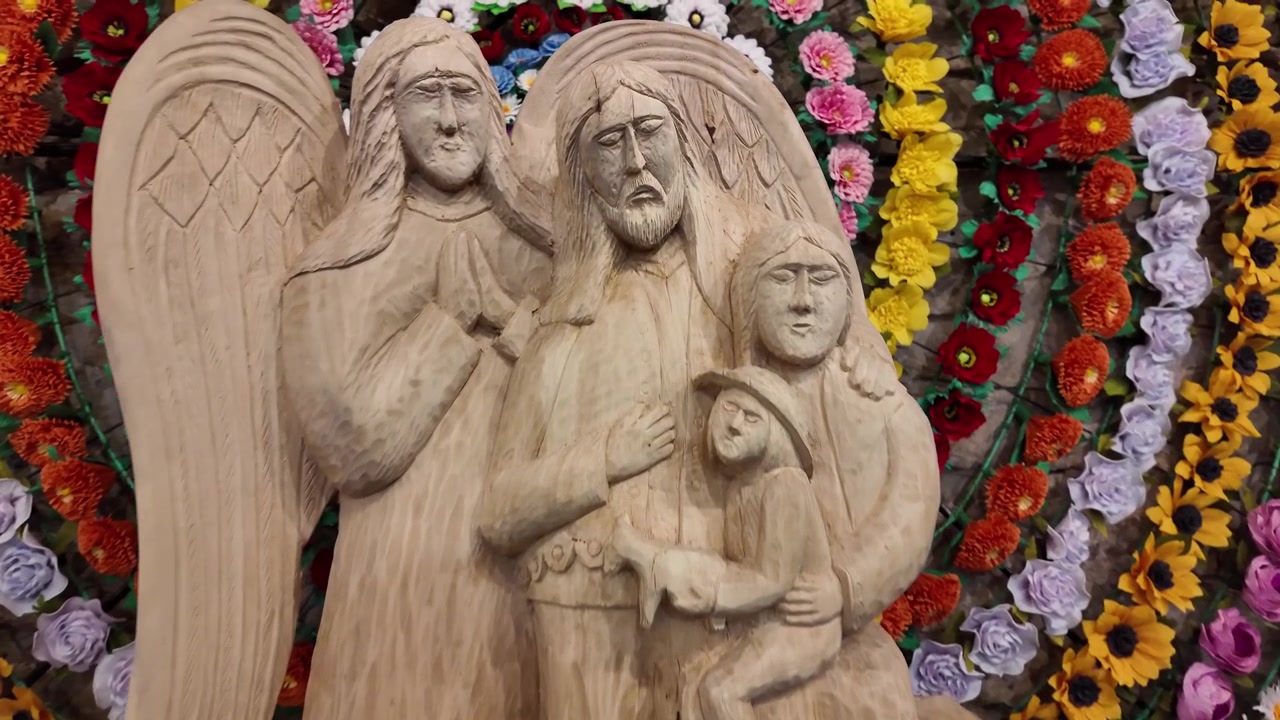 Light‑wood figurine of an angel protecting a three‑person family. Colorful two‑meter floral circle forms a striking museum backdrop.