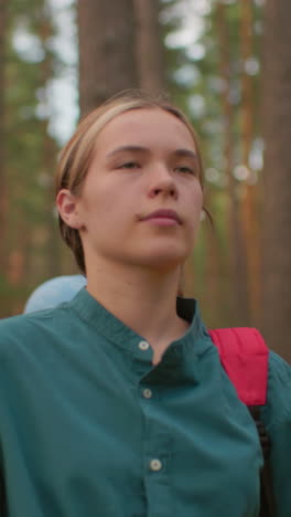 dama con mochila roja y camisa verde camina a través de un sendero del bosque sereno, rodeado de árboles altos y naturaleza pacífica, su amiga, con una bufanda azul, camina detrás de ella, mirando hacia abajo en pensamiento