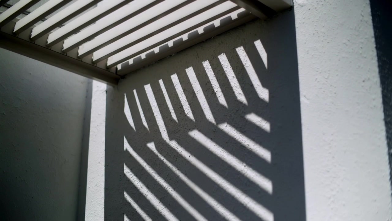 Ângulo do sol variável projetando sombras diagonais que deslizam pela parede de estuque sob um beiral de canto ripado.