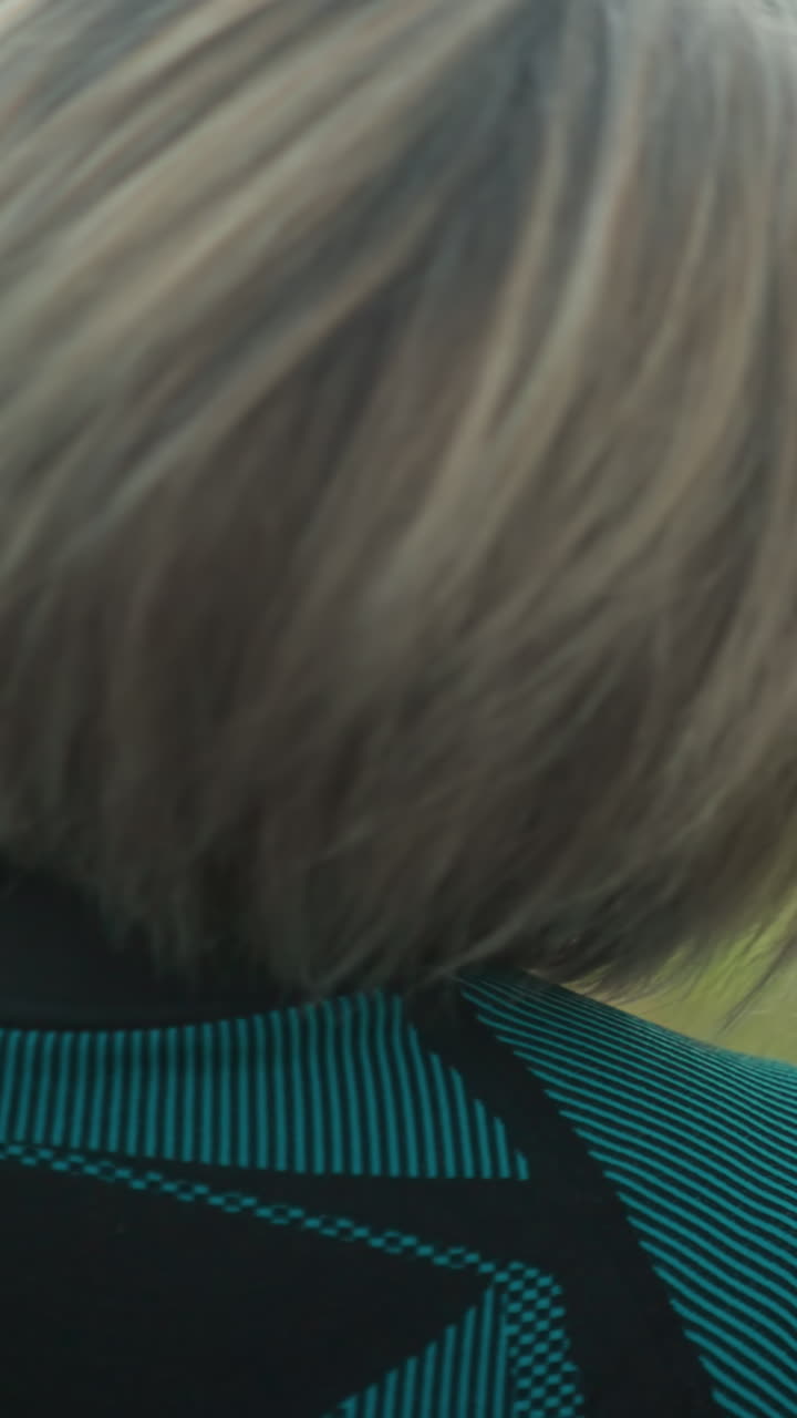 Back view of woman in black and green suit, eyes closed, tilting her head slowly from right to left, stretching and relaxing her neck muscles outdoors in a peaceful grassy field under a cloudy sky