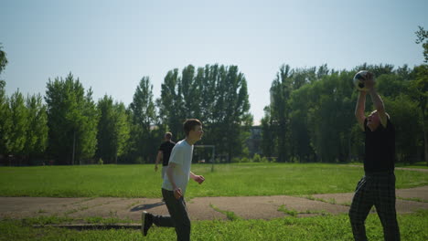 a grandfather and grandson play football outdoors, the grandfather throws, the grandson heads the ball back, there's another ball nearby, trees, and a goalpost in the distance