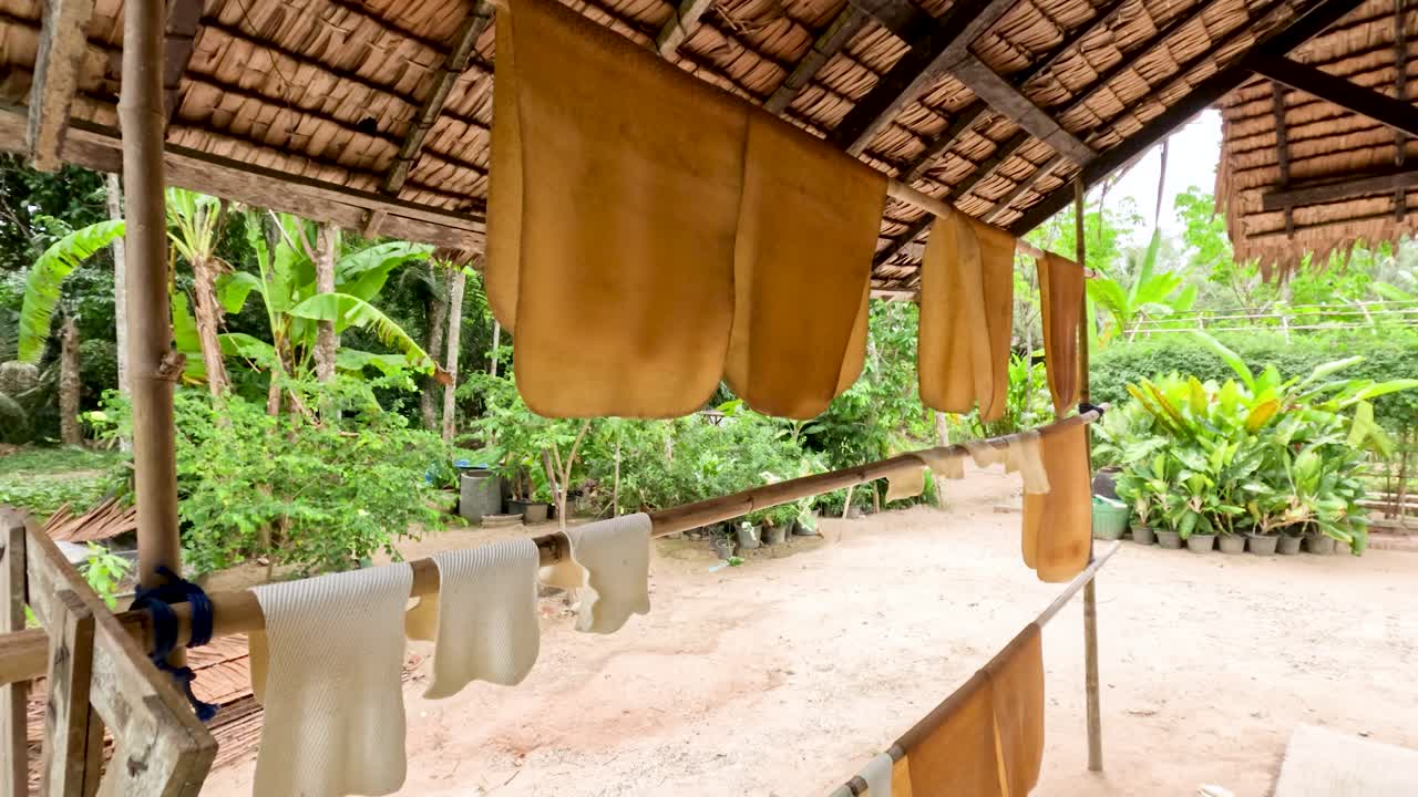 Natural rubber sheets hang drying under a rustic shelter, surrounded by tropical greenery and daylight