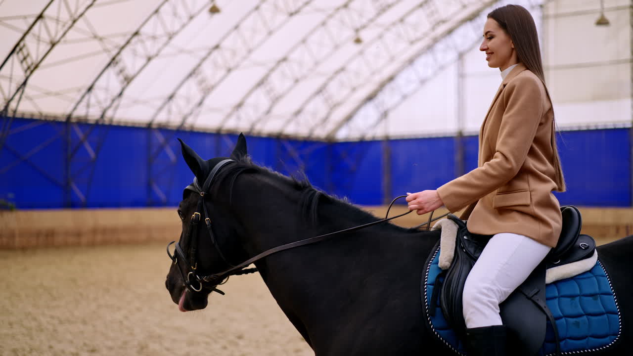 Happy smiling lady taking lessons of horse riding. Beautiful long-haired brunette rides on a black horse slowly. Manege backdrop.