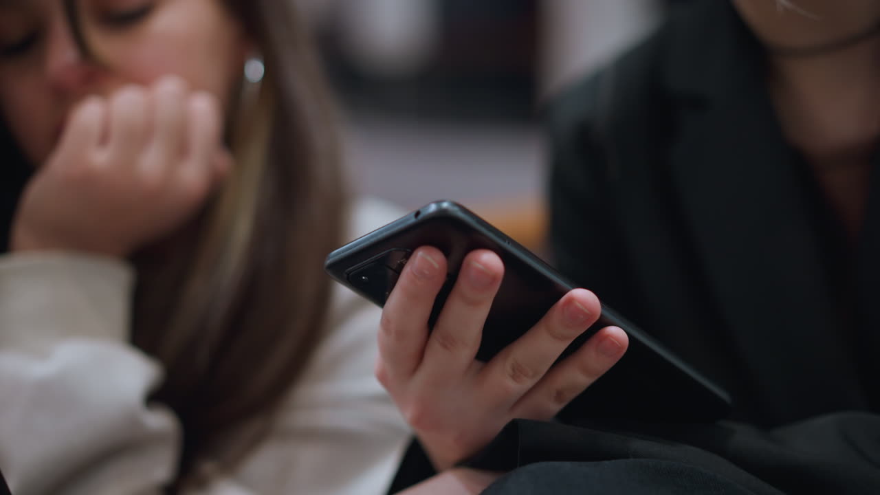 Close up hand view of young woman operating smartphone while another person appears blurred in background showing technology use