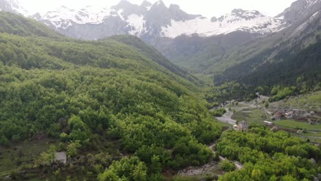 Drone-view-in-Albania-flying-in-the-alps-showing-green-forest-on-a-valley-surrounded-by-mountain-with-snowy-peaks-in-Valbon?