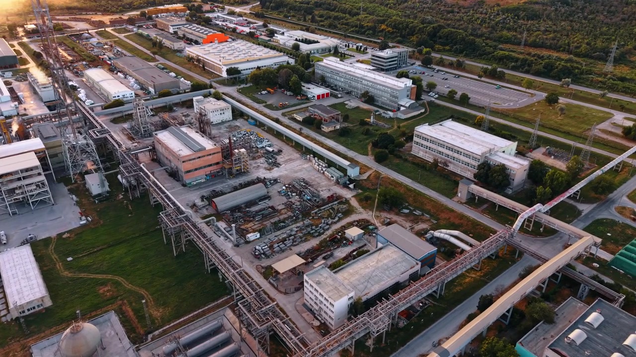 Pipeline network connecting buildings in an industrial complex. High angle drone view shows elevated pipes, processing structures, service roads, offices and surrounding vegetation.