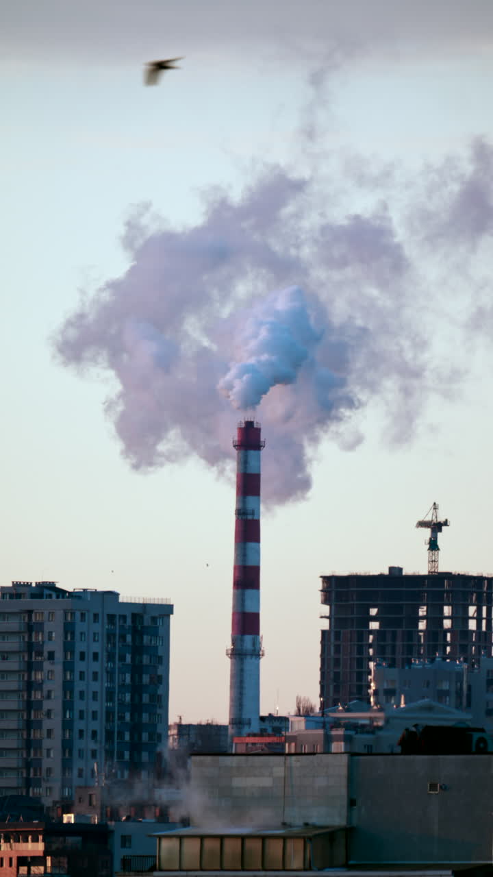 Ciminiera industriale che rilascia fumo denso nel cielo sopra un cantiere edile cittadino. Verticale.